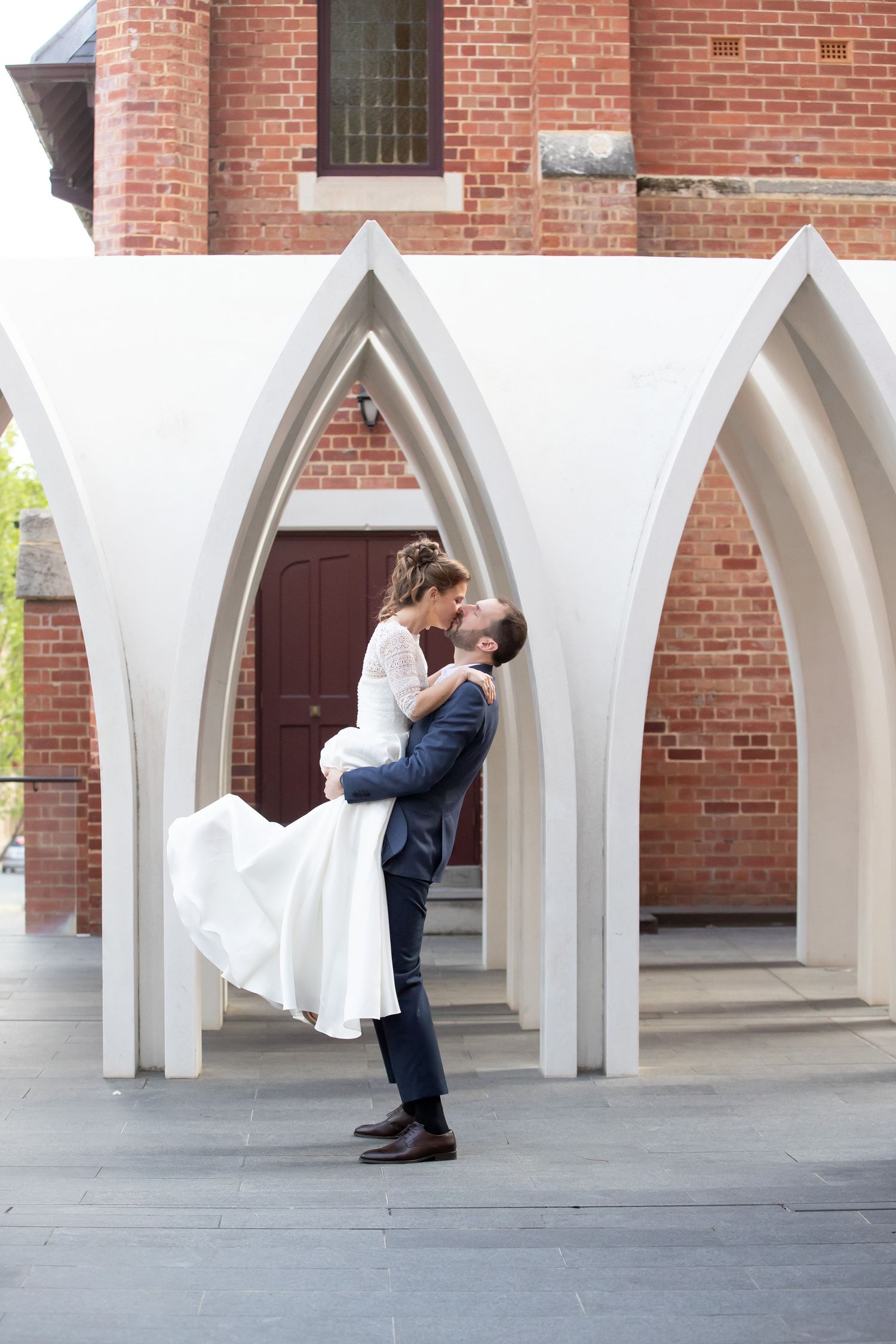 Groom lifting bride in white lace dress among white gothic arches at a brick church.