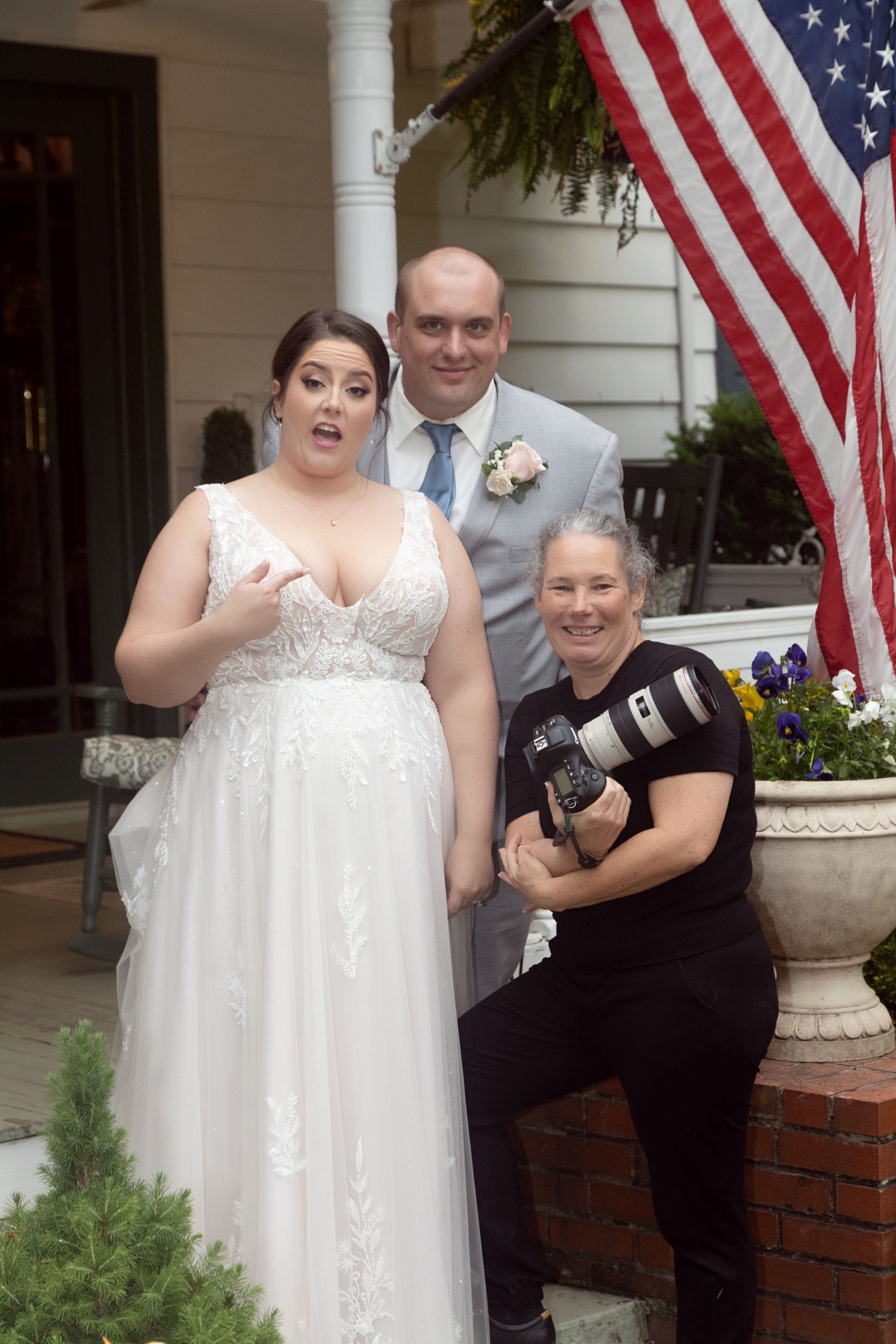 Bride in white gown and groom in gray suit pose with smiling photographer holding camera near American flag.