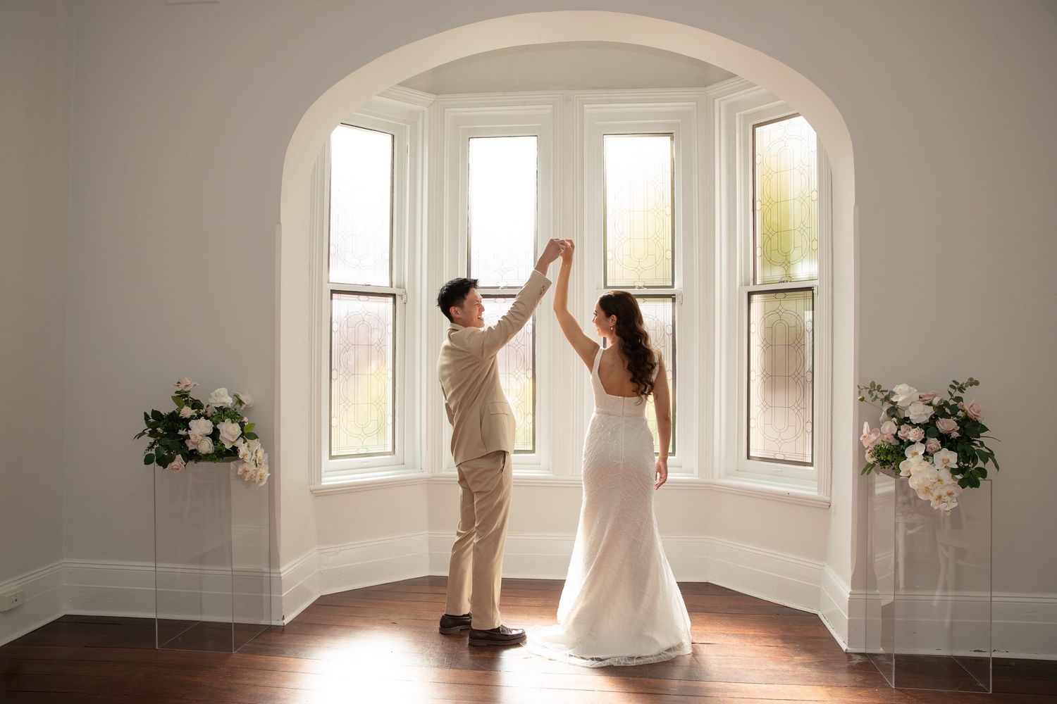 A couple dances elegantly near arched windows in a bright white room decorated with floral arrangements.