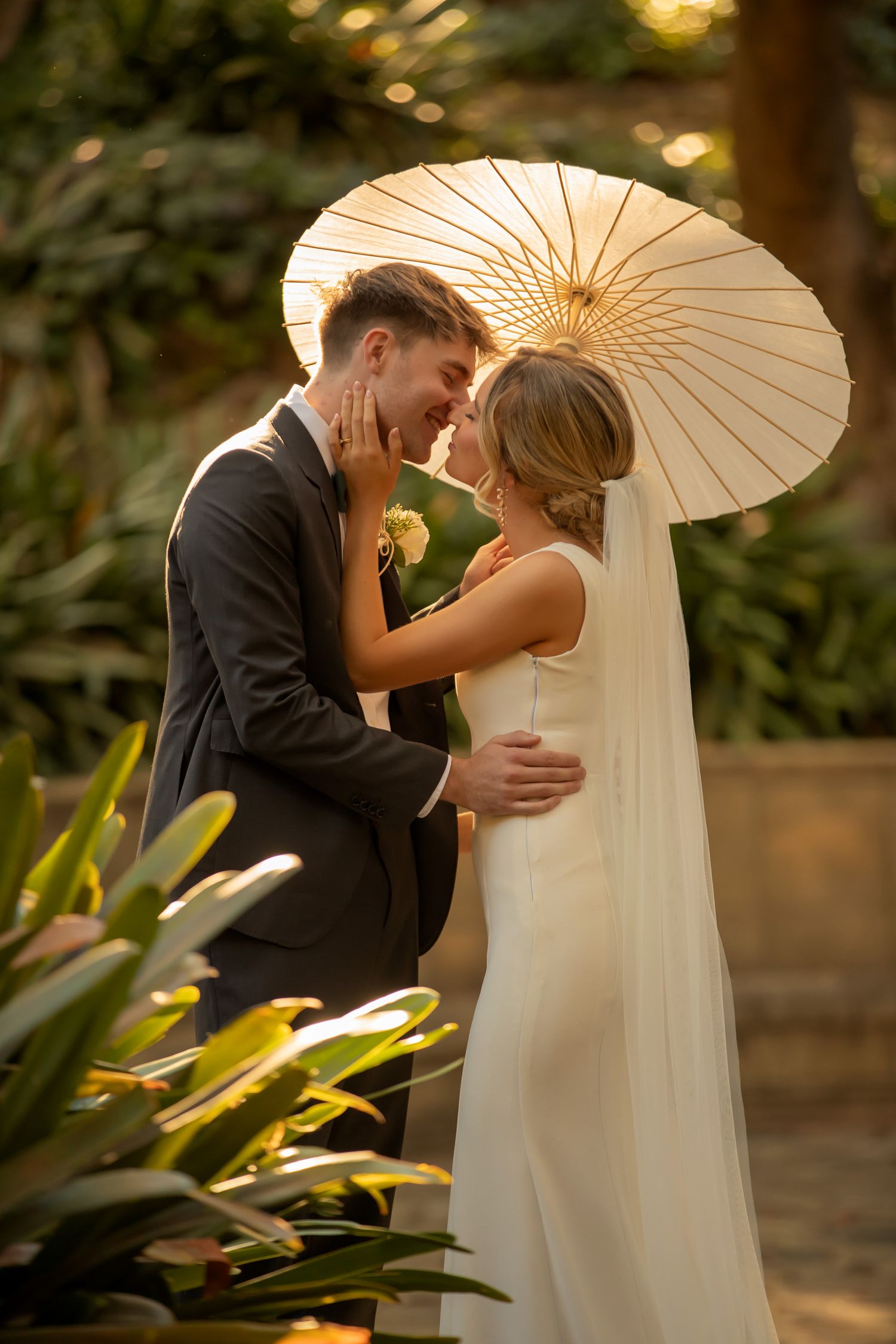 Bride and groom share a romantic moment under a white parasol umbrella, surrounded by lush greenery in warm golden light.