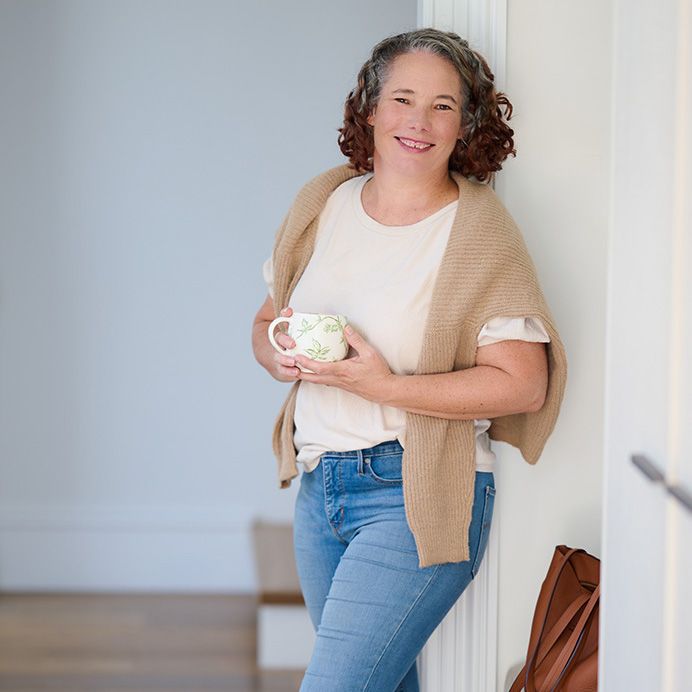Smiling woman with curly hair holding a floral mug, wearing jeans and a tan wrap, leaning against a white wall.