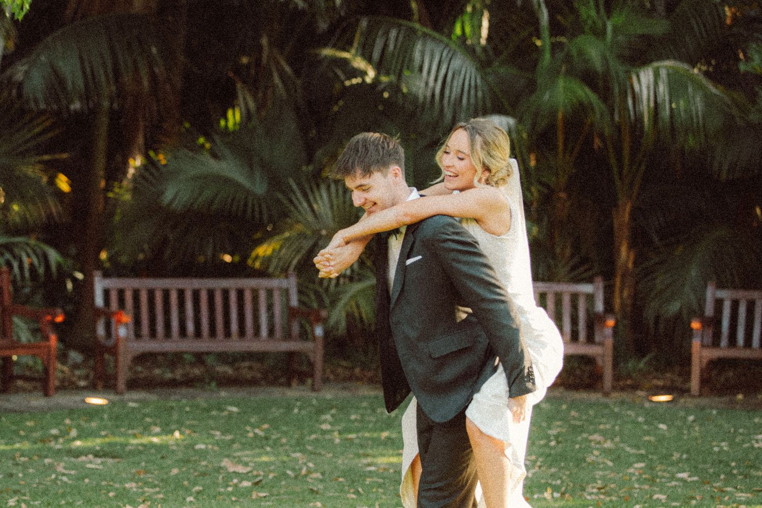 Bride in white dress joyfully hugging groom in black suit outdoors with lush tropical greenery and wooden bench.
