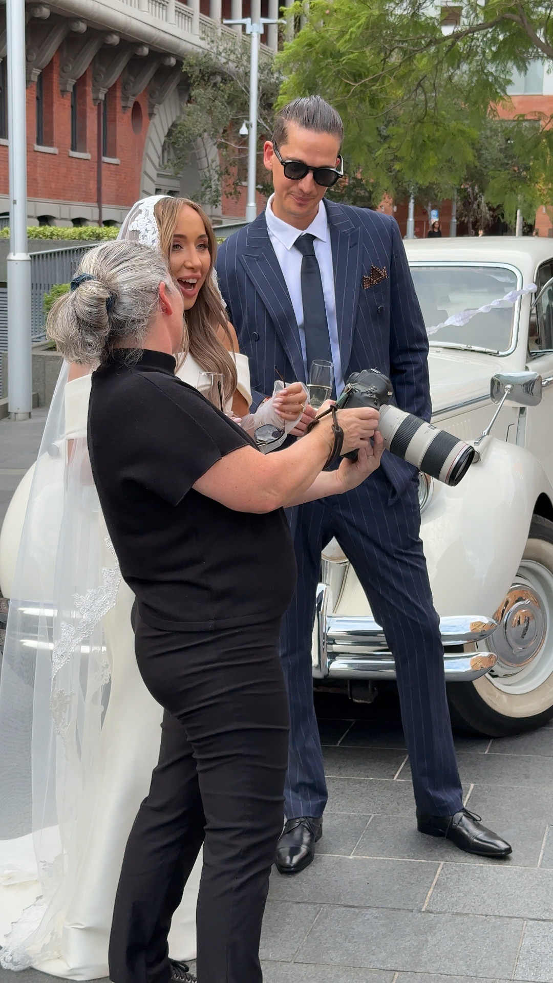 Wedding photographer shows camera to bride in white gown and groom in navy pinstripe suit beside vintage white car.