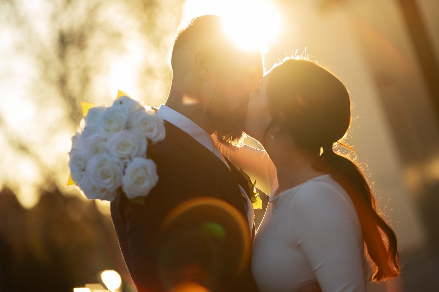 Bride and groom sharing a romantic kiss at sunset with golden light and white rose bouquet.