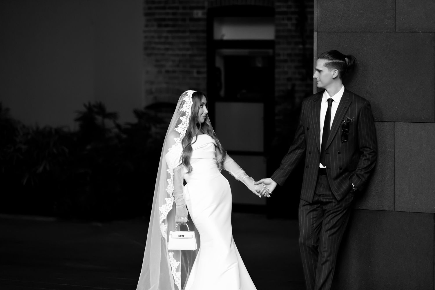 Black and white wedding photo of bride in veil and groom in suit holding hands outdoors.
