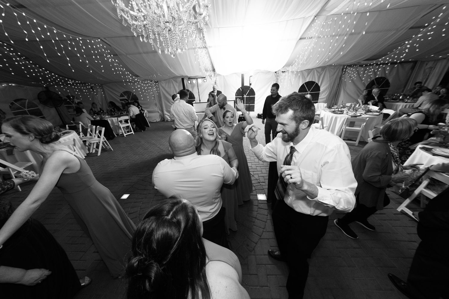 Black and white photo of guests dancing and celebrating at an elegant wedding reception under a decorated tent.