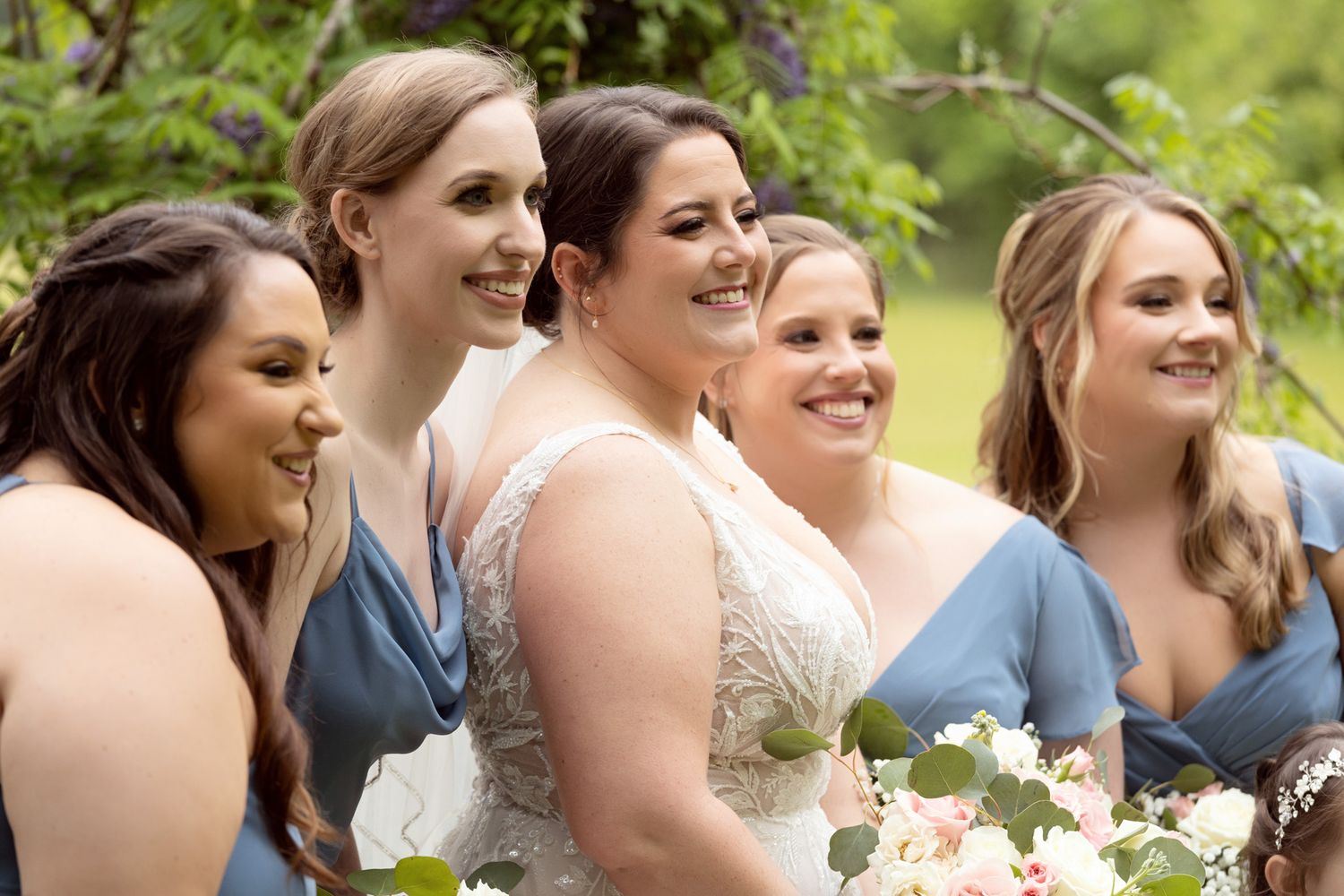 Bride in white lace gown smiles with bridesmaids in dusty blue dresses, holding floral bouquet outdoors.