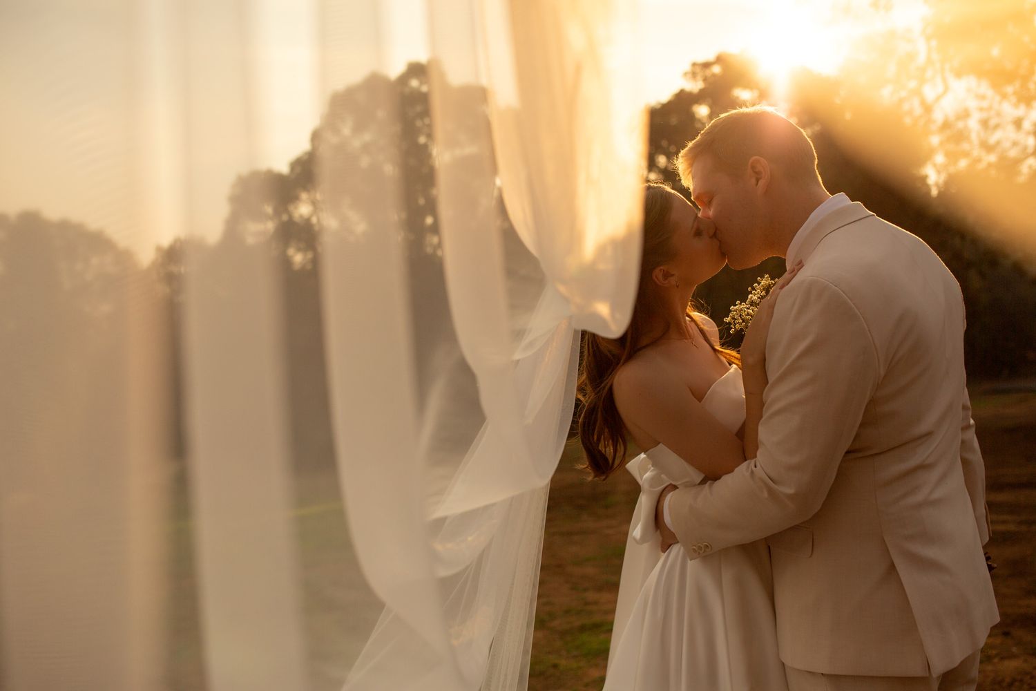 Couple sharing a romantic kiss at golden hour, bride in white gown with veil, groom in beige suit.