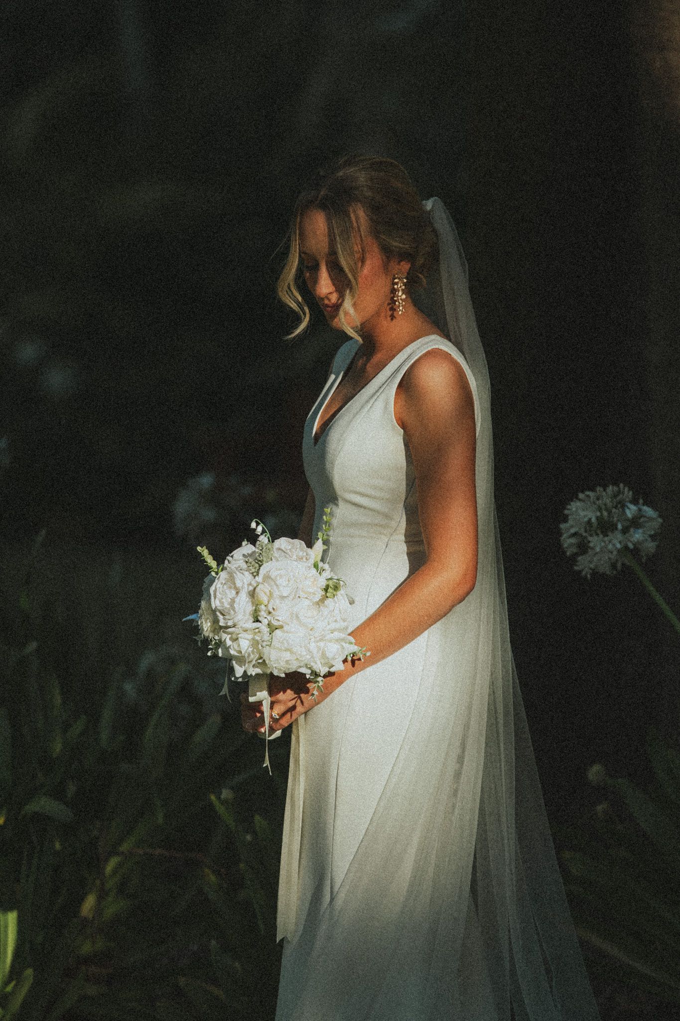 Bride in white gown and veil holds white floral bouquet, looking down in a dark garden setting.