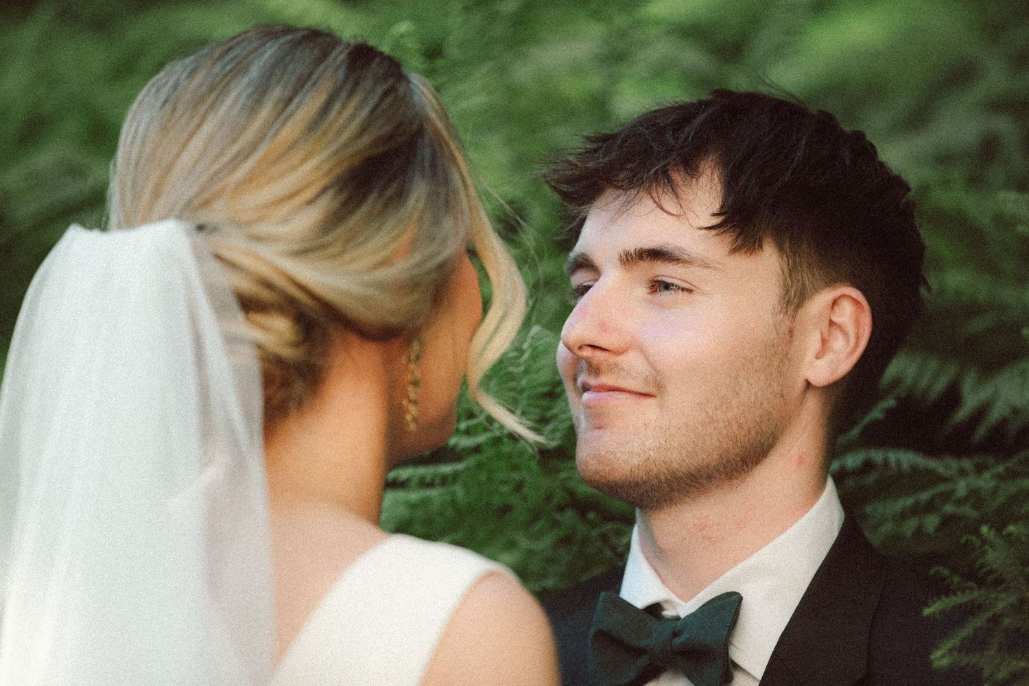 Groom in black tuxedo gazes lovingly at bride wearing veil, surrounded by lush green ferns outdoors.