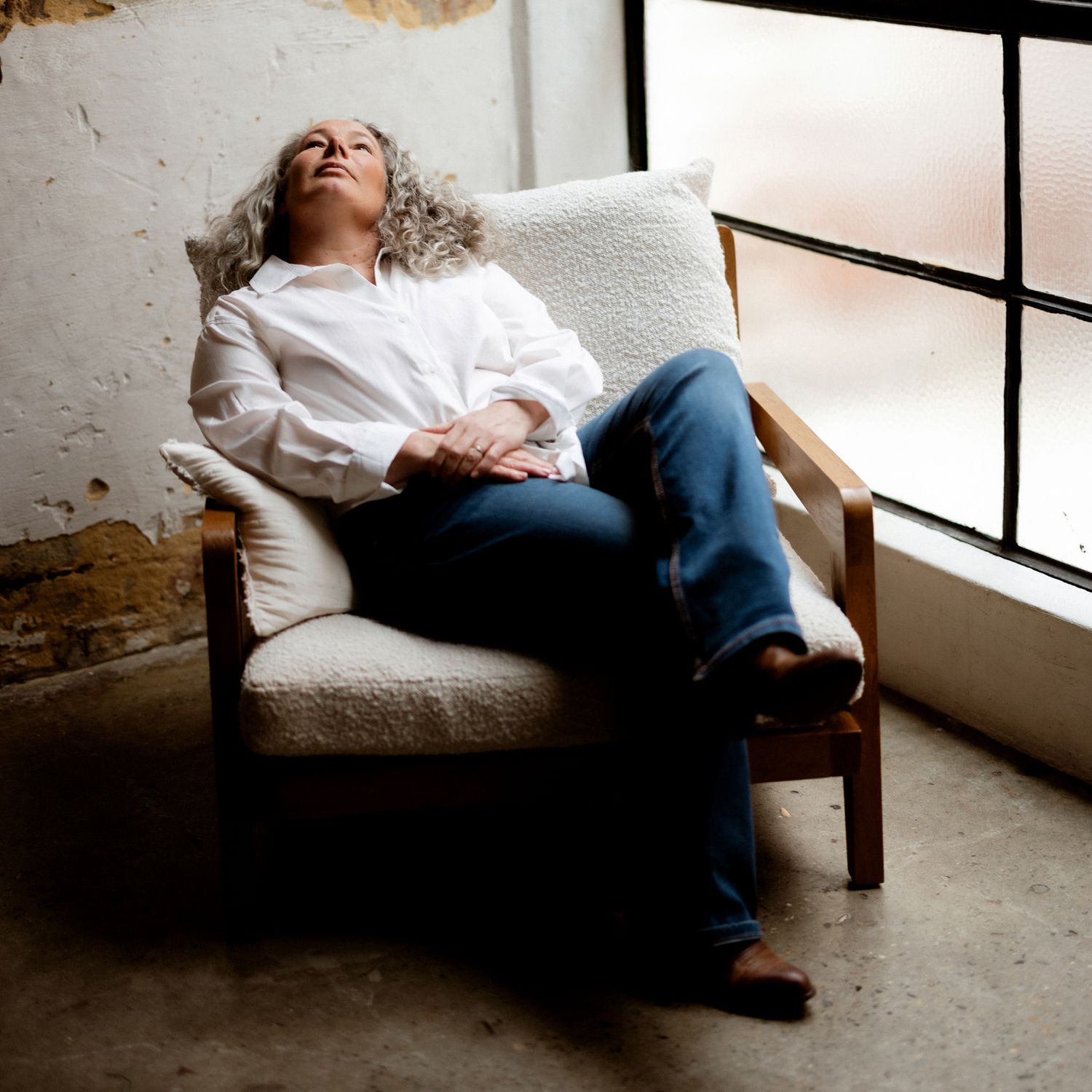 A relaxed person with gray hair reclines in a white armchair near a window, wearing a white shirt and jeans.