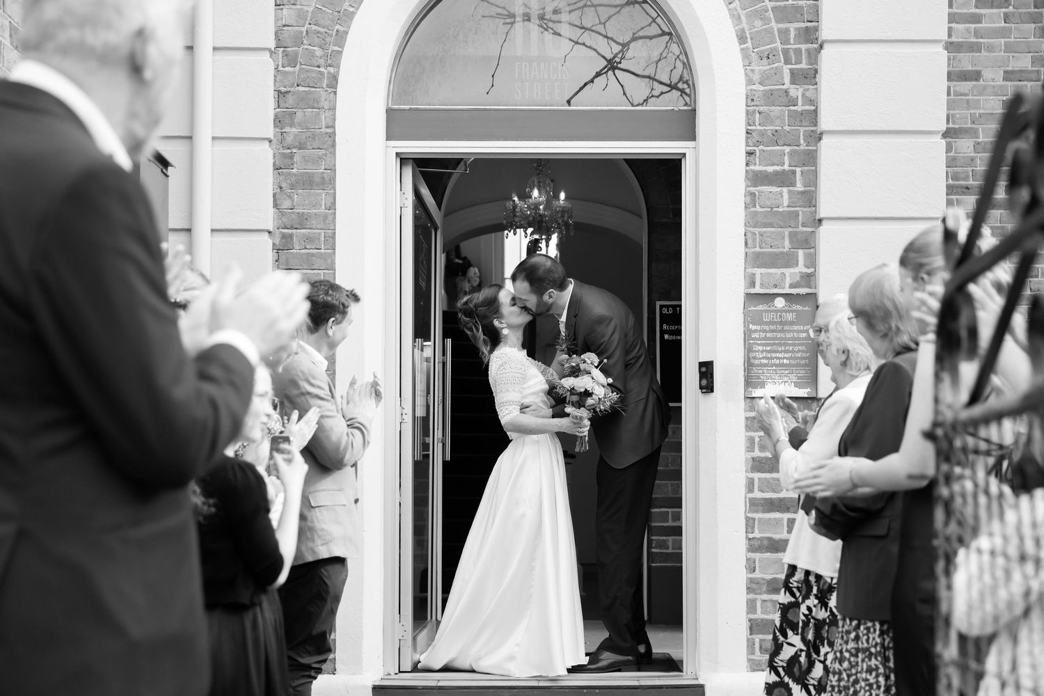 Newlyweds share a romantic kiss in a brick building doorway as wedding guests look on in black and white.