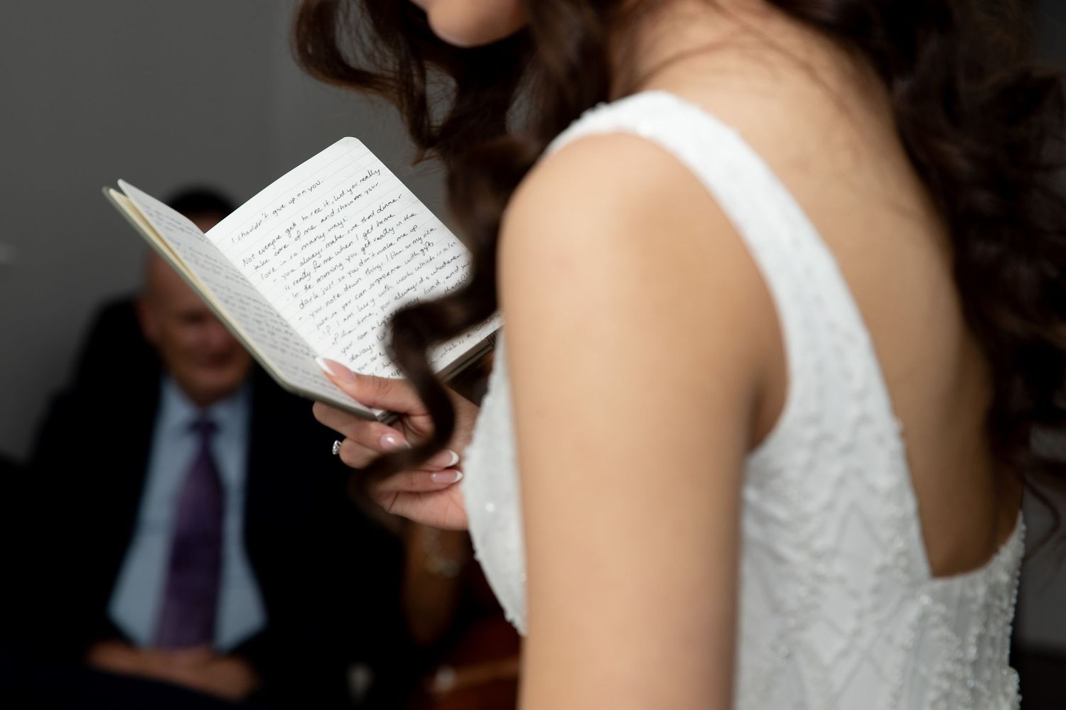 Bride in a white sleeveless gown reading handwritten wedding vows from a small notebook during the ceremony.