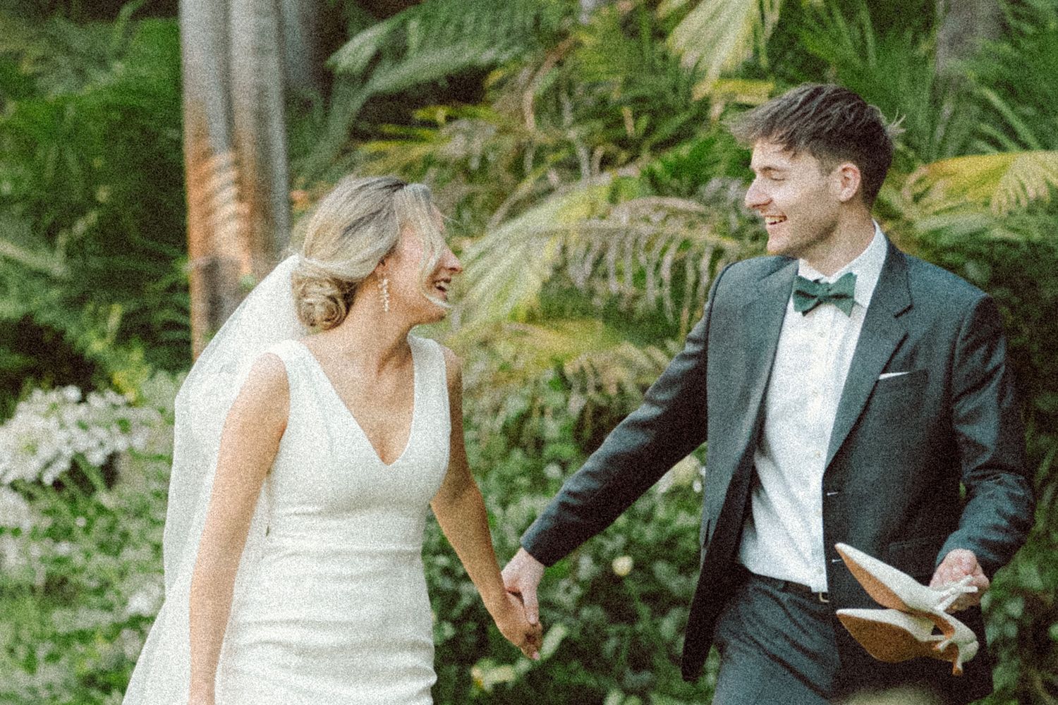 A laughing bride in white gown and groom in dark suit with bow tie share a joyful moment outdoors amid lush tropical greenery.