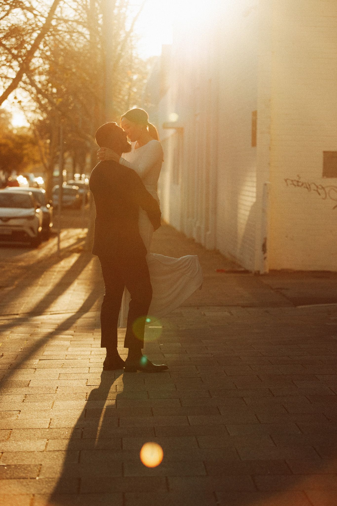 A couple shares a romantic embrace on a sunlit city sidewalk during golden hour, with warm lens flare and long shadows.