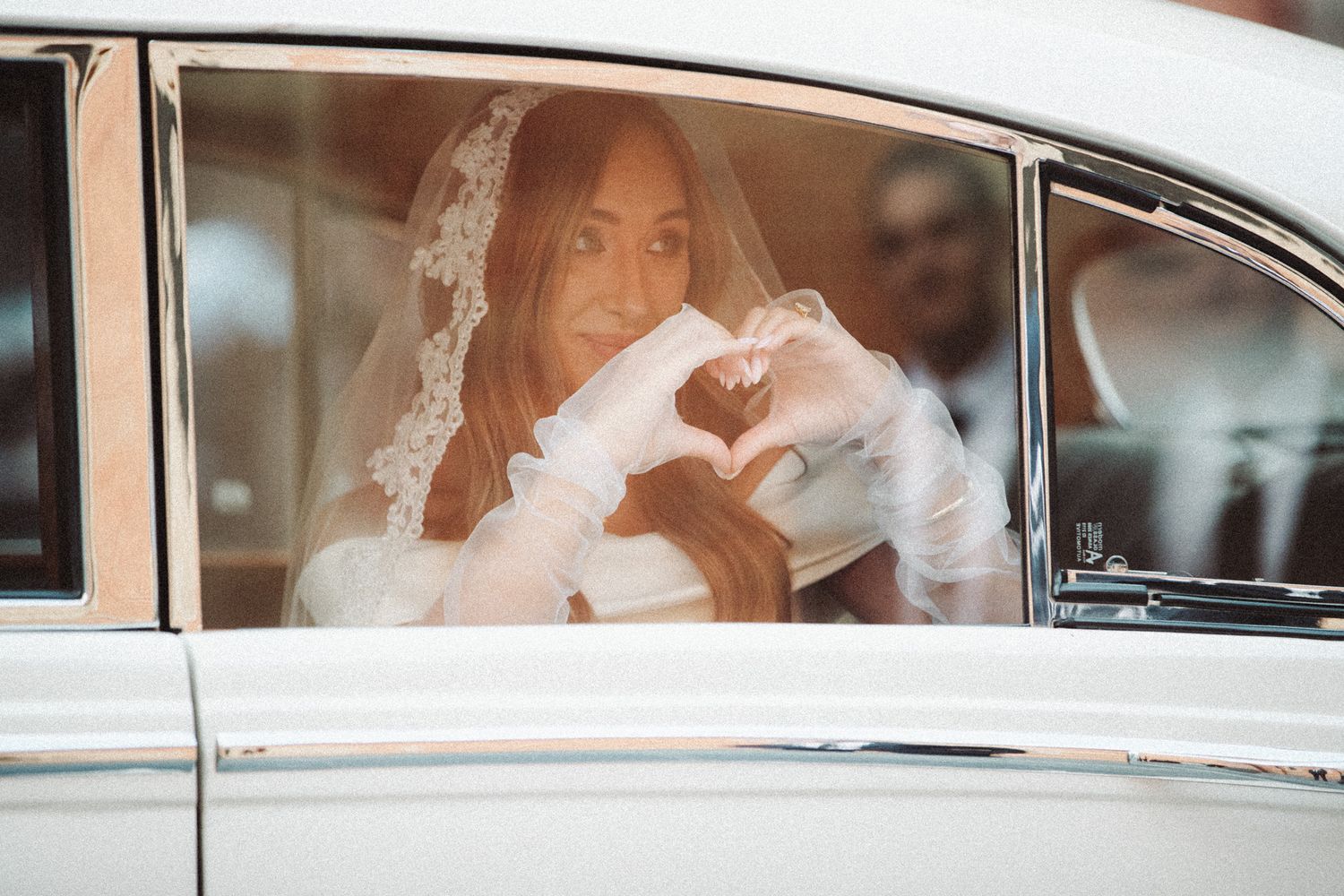Bride in white veil and gloves makes heart shape with hands through vintage car window on wedding day.