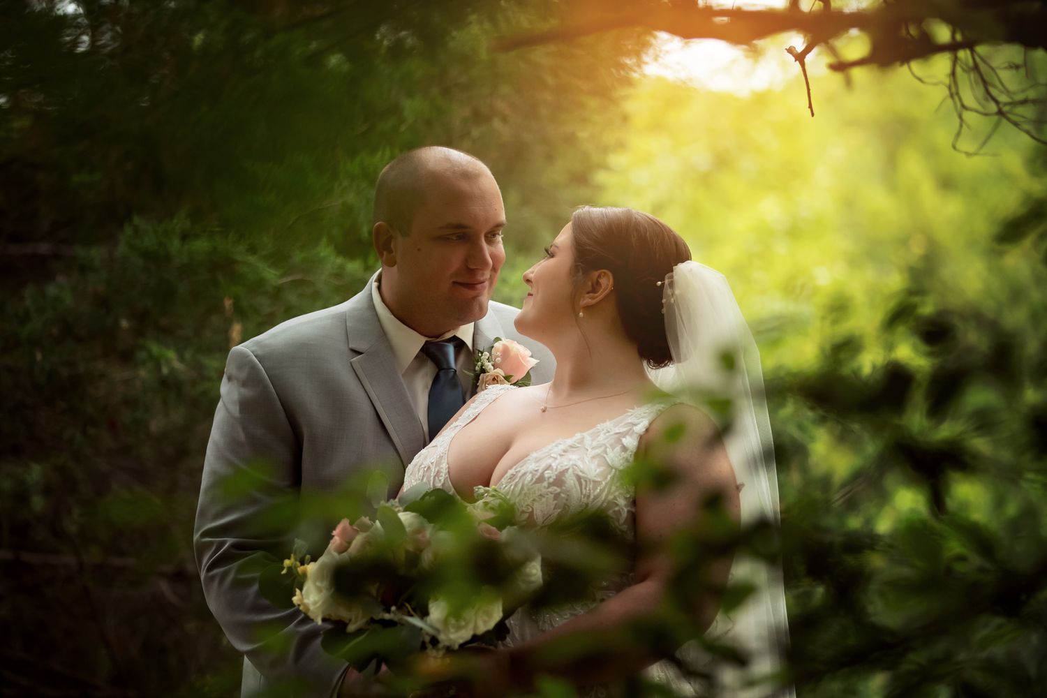 A bride and groom share a romantic moment surrounded by lush green foliage in golden sunlight.