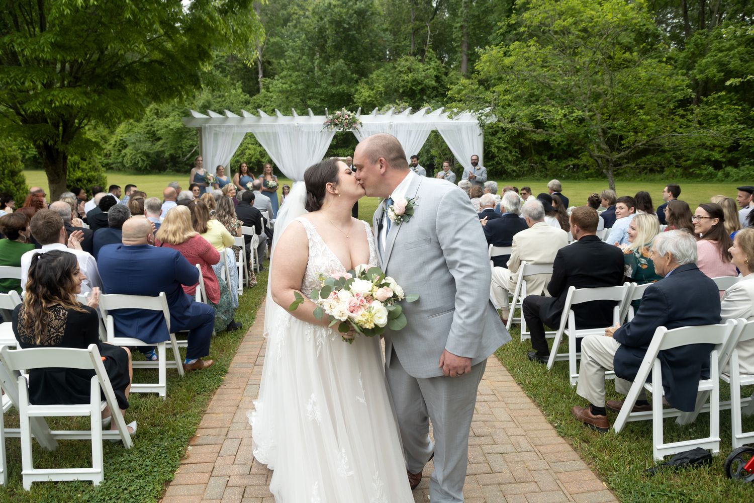 Bride and groom share a kiss at outdoor wedding ceremony with guests seated on white chairs amid lush greenery.
