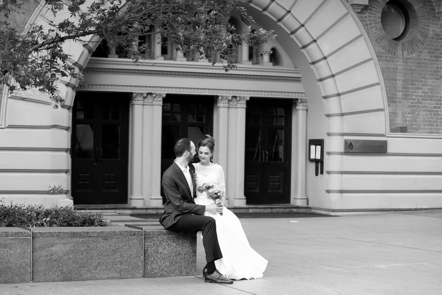 Black and white wedding photo of bride and groom sitting together outside an elegant arched building.