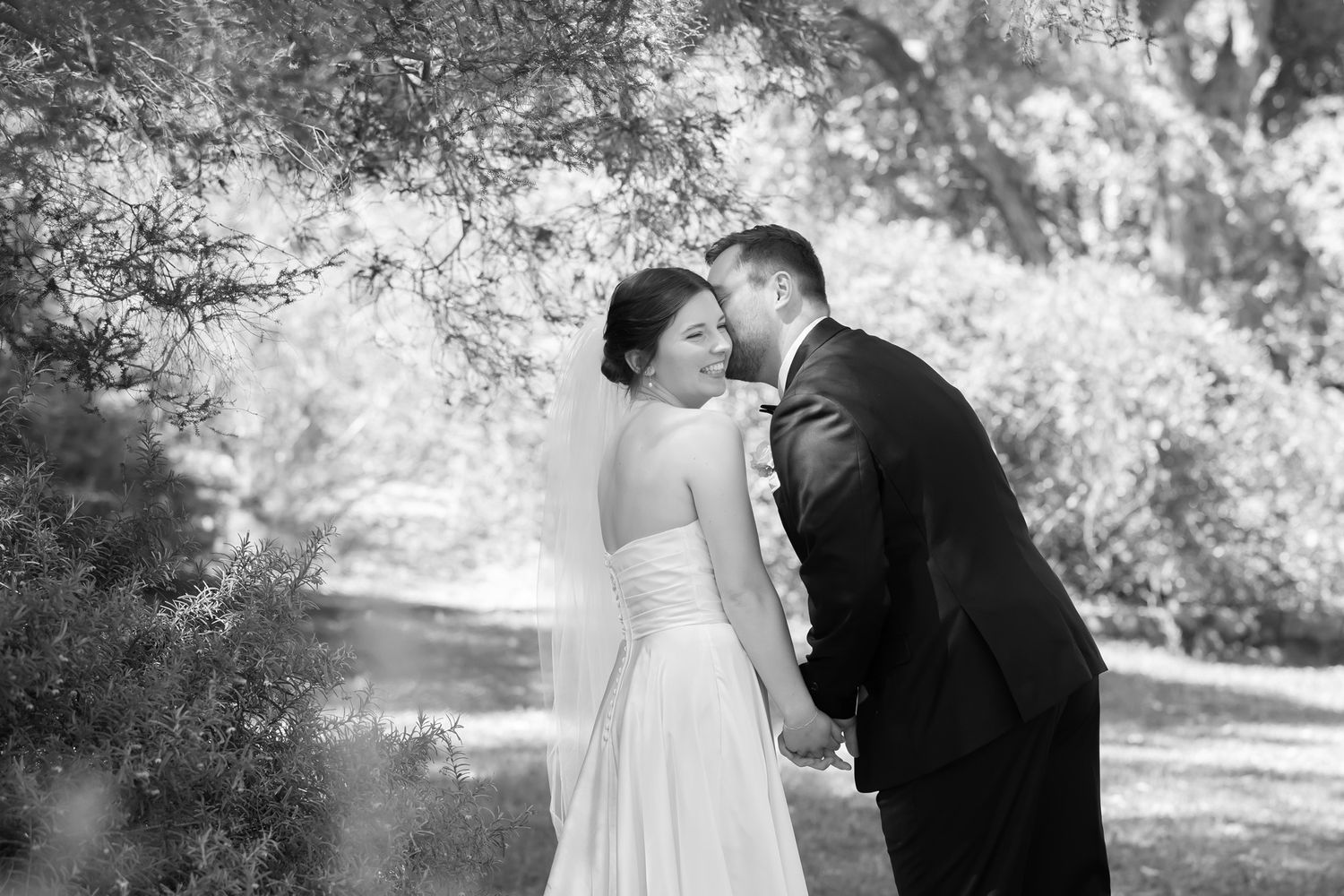 Black and white photo of bride and groom sharing a romantic moment under lush trees in a garden setting.