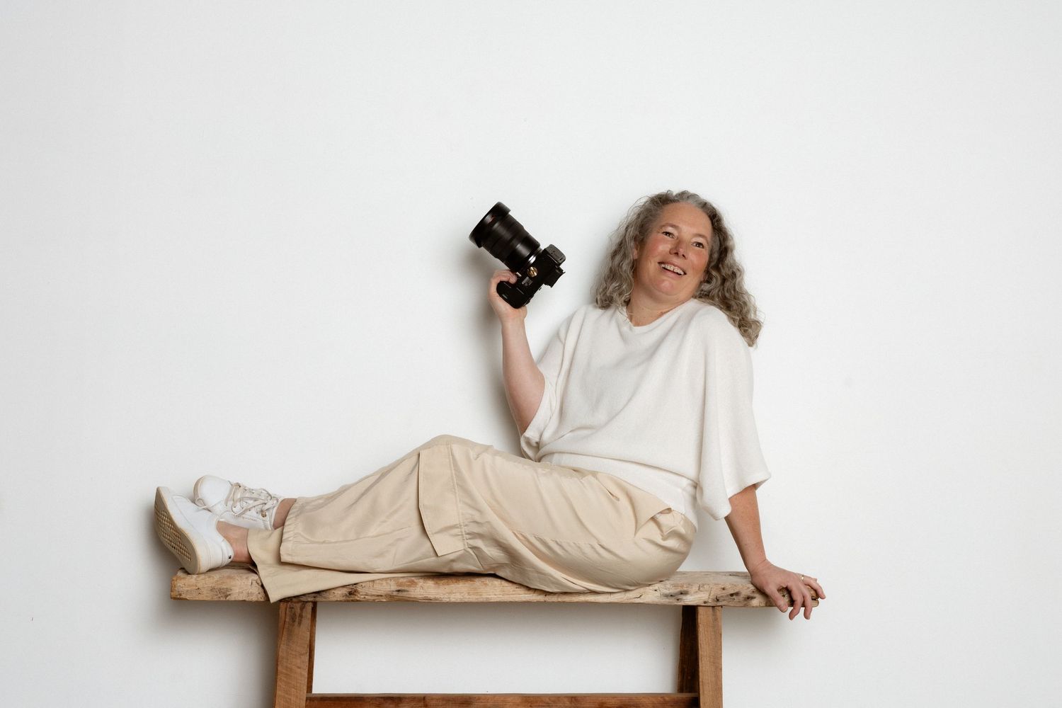 Smiling woman with curly hair holding binoculars while relaxing on a wooden bench against a white background.