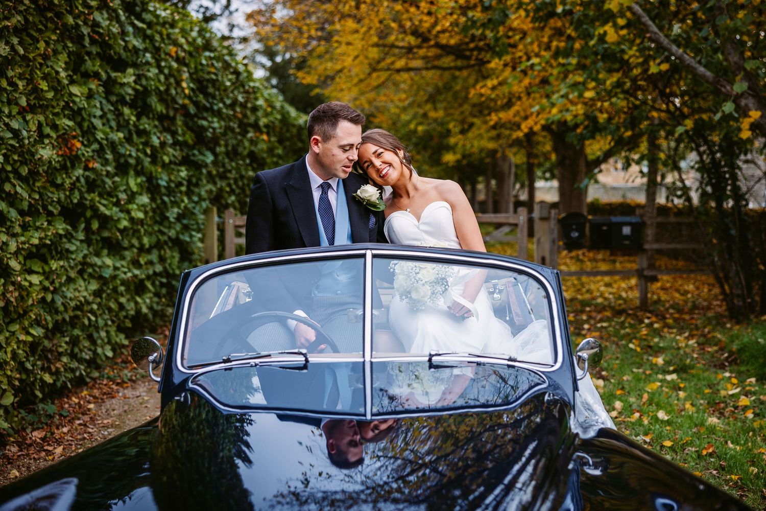 Newlyweds share a romantic moment in a vintage blue convertible car surrounded by autumn foliage.