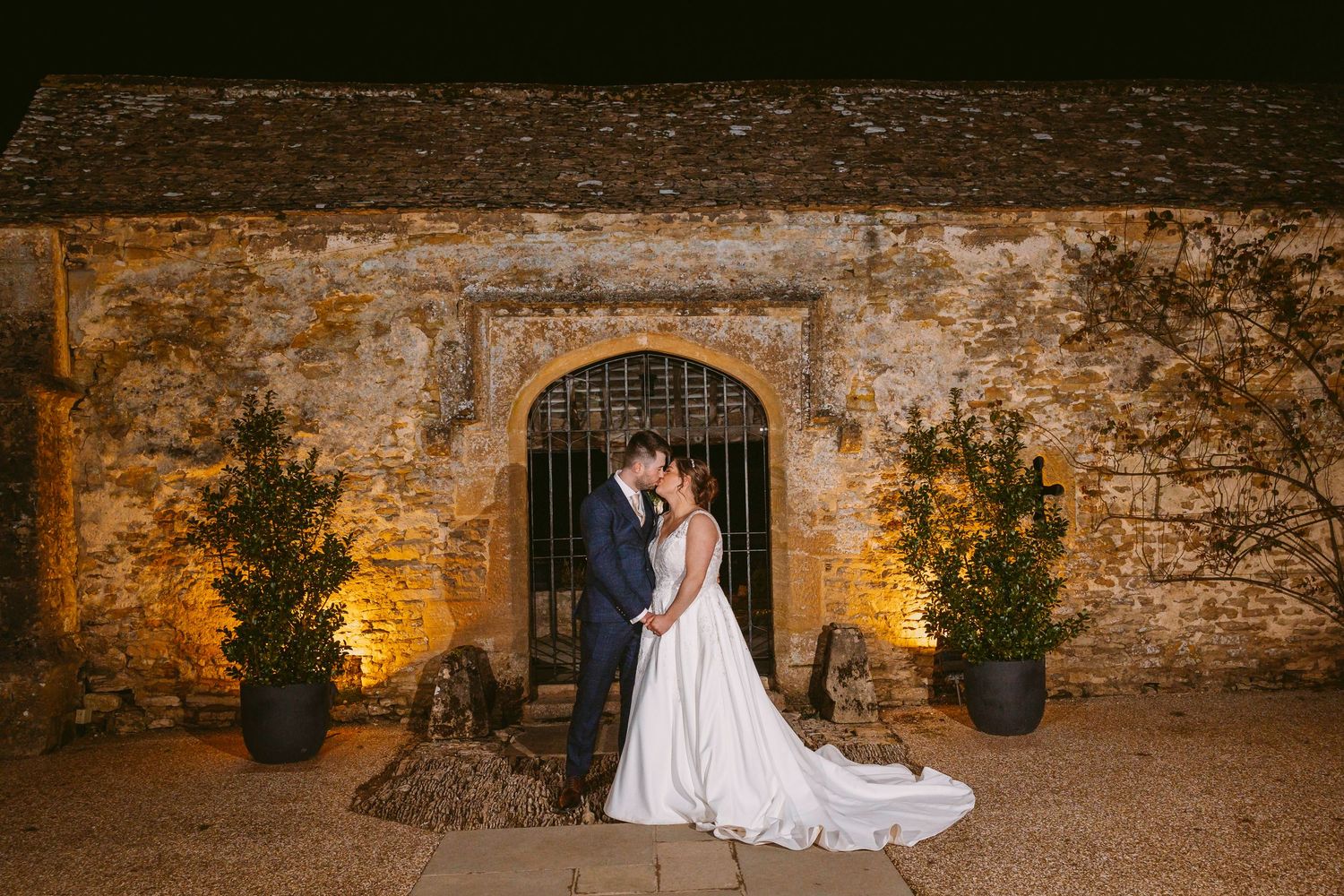 A romantic evening scene outside a stone archway with warm lighting and potted trees at an elegant venue.