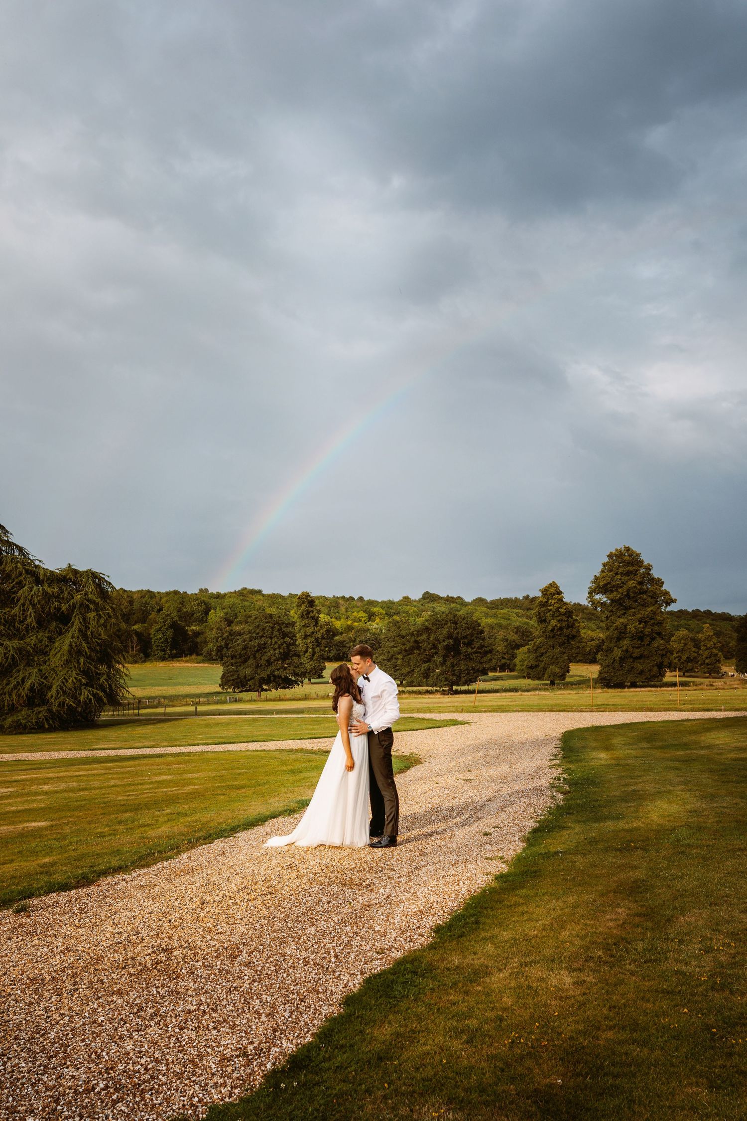 Wedding couple standing on gravel path with dramatic stormy sky and rainbow.