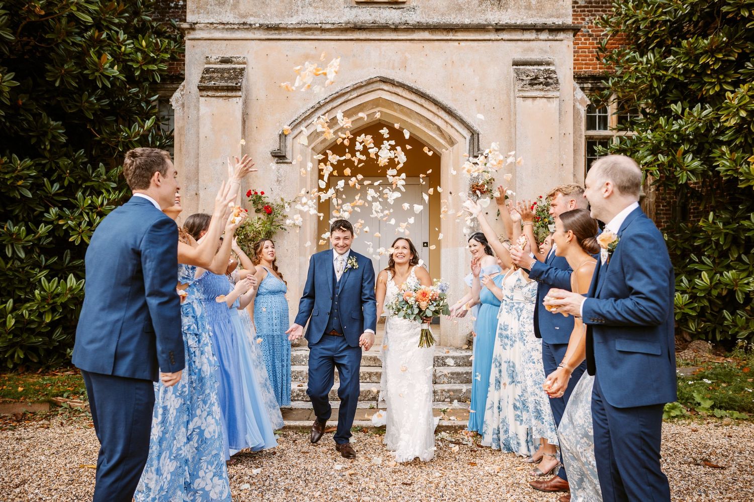Joyful wedding party throws confetti at a historic stone archway entrance.
