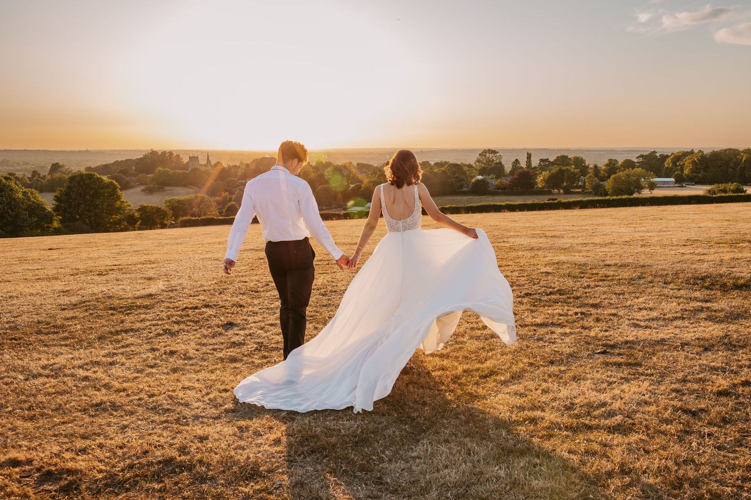 Bride and Groom couple in wedding attire walk together on a hilltop during golden hour sunset.