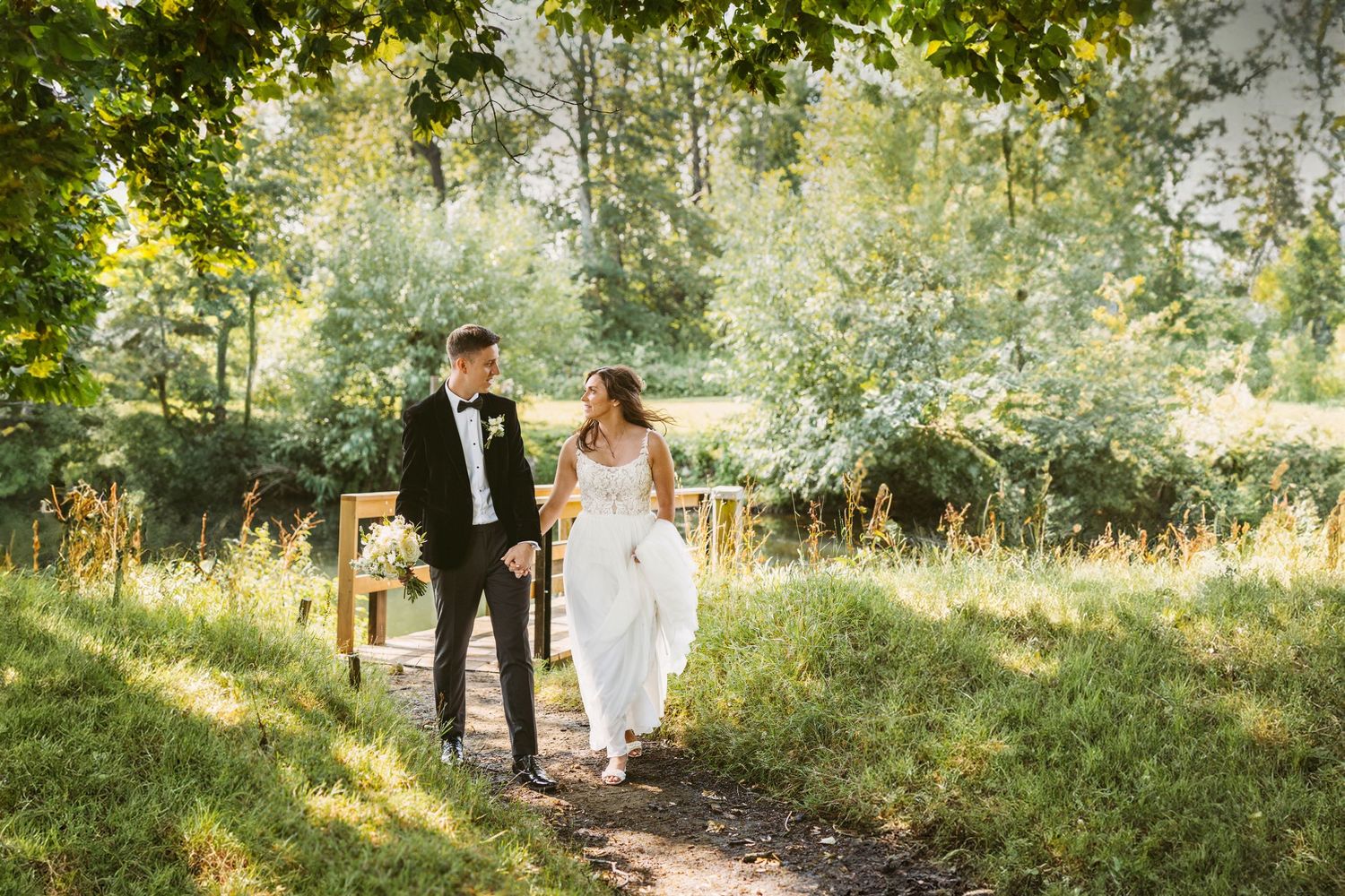 A wedding couple strolls through a sunlit woodland path in formal attire.