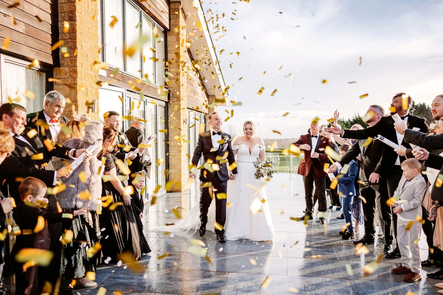 Wedding guests toss flower petals in celebration as newlyweds walk through a covered outdoor corridor.