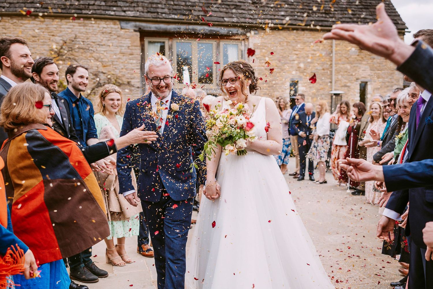 Newlyweds walk through blue confetti being thrown by wedding guests outside a rustic brick venue.