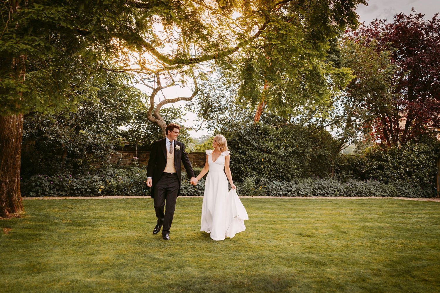 A couple walks together through a lush garden setting during a sunset wedding celebration.