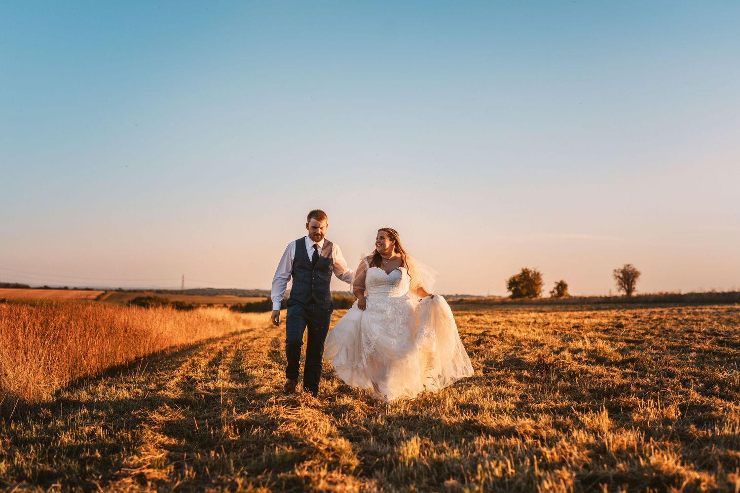 Bride and groom walking hand in hand through a golden field at sunset Stone Barn.