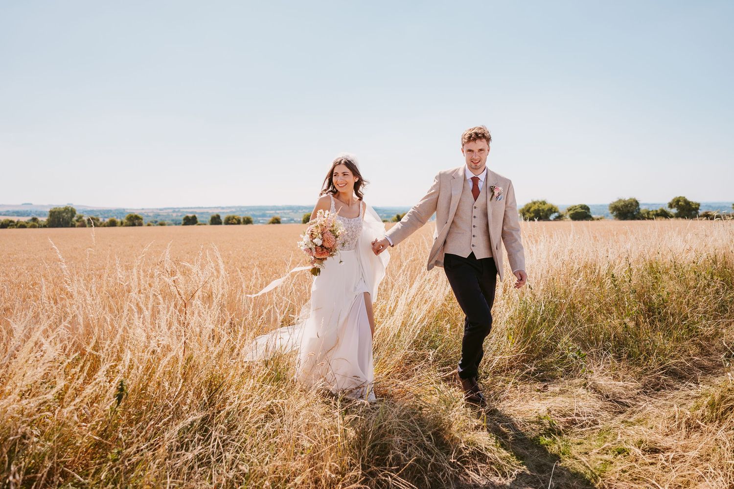 A Bride and Groom couple walks hand in hand through a golden wheat field on a sunny summer afternoon.