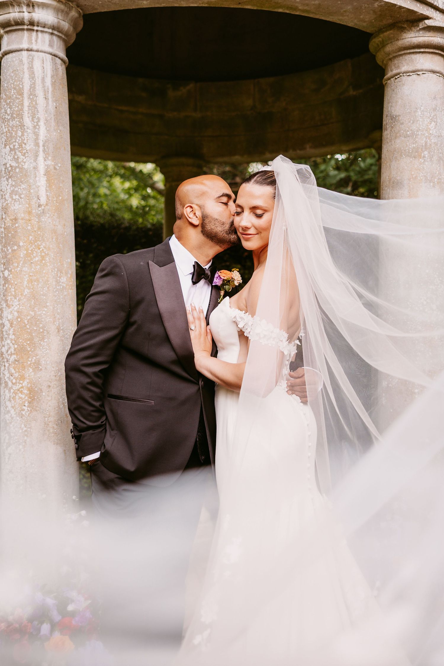 A romantic wedding moment captured under a stone archway with flowing white veil and formal attire.