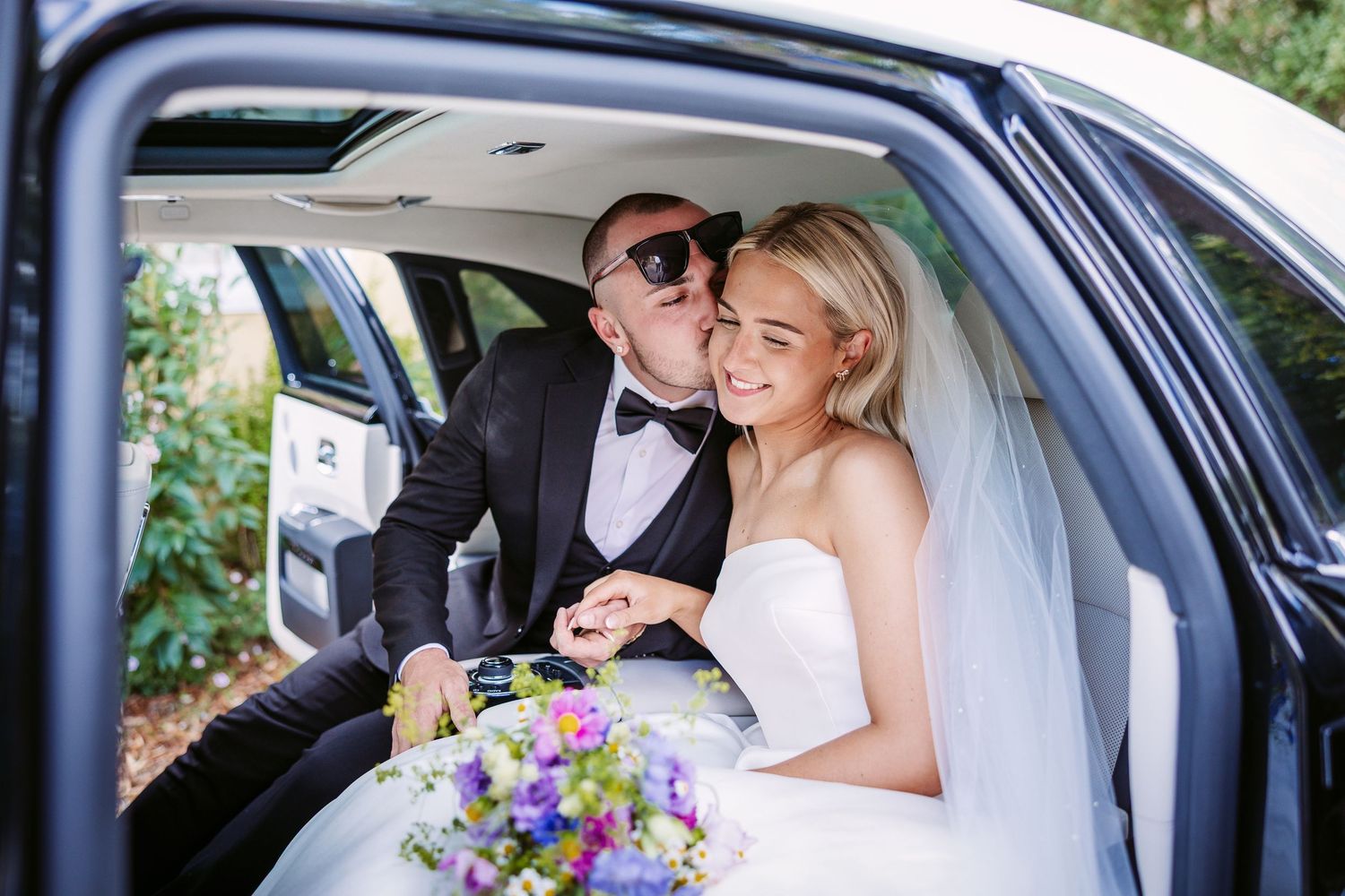 A newlywed couple sits together in the backseat of a car with a colourful purple and white wedding bouquet.