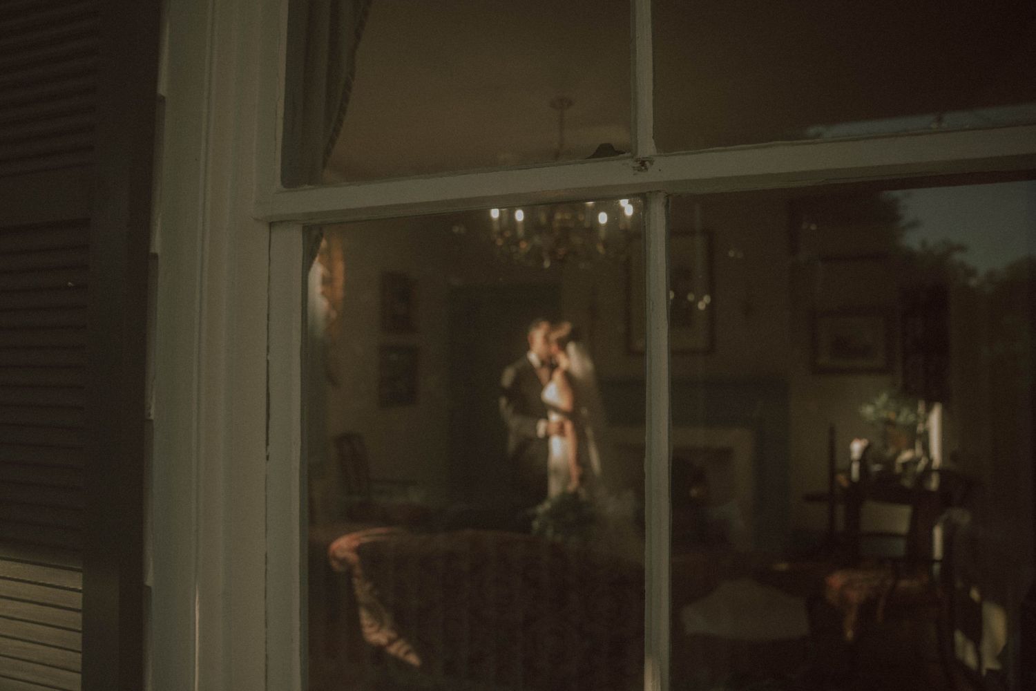 Through a window at night, a silhouette stands illuminated in a cozy living room interior.