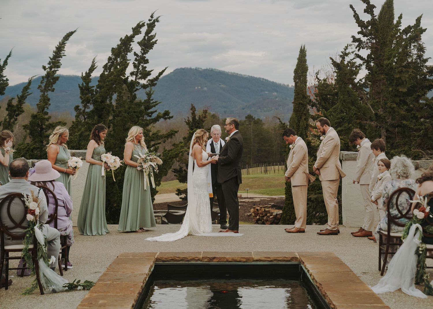 An outdoor wedding ceremony takes place on a mountaintop setting with a reflecting pool and evergreen trees in the background.