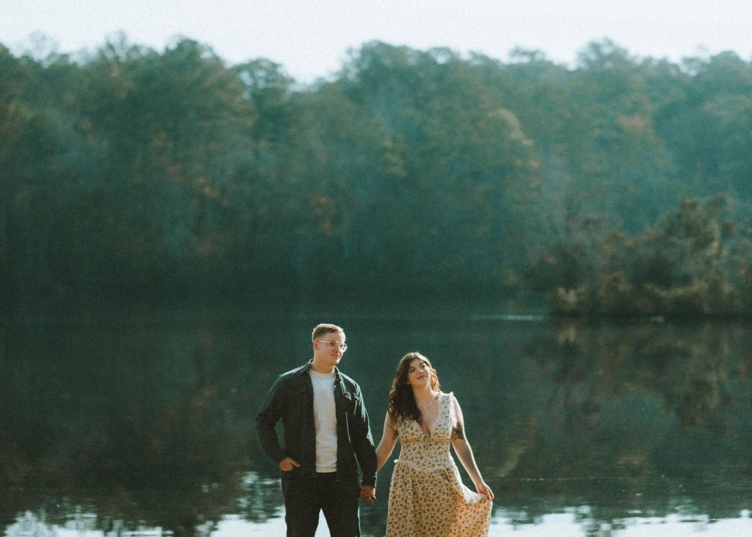 A couple walks hand in hand along a serene lake path surrounded by lush greenery and autumn trees.