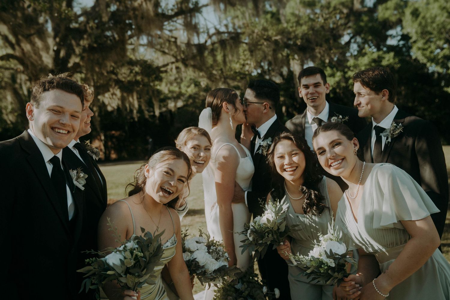 A candid wedding party moment outdoors with bridesmaids and groomsmen laughing together against a backdrop of trees.