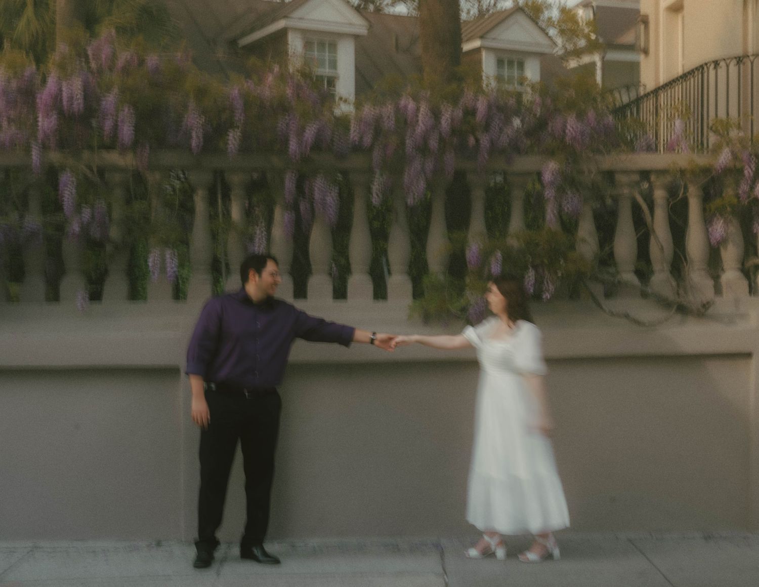 A couple shares a romantic moment on a path beneath blooming purple wisteria vines at sunset.