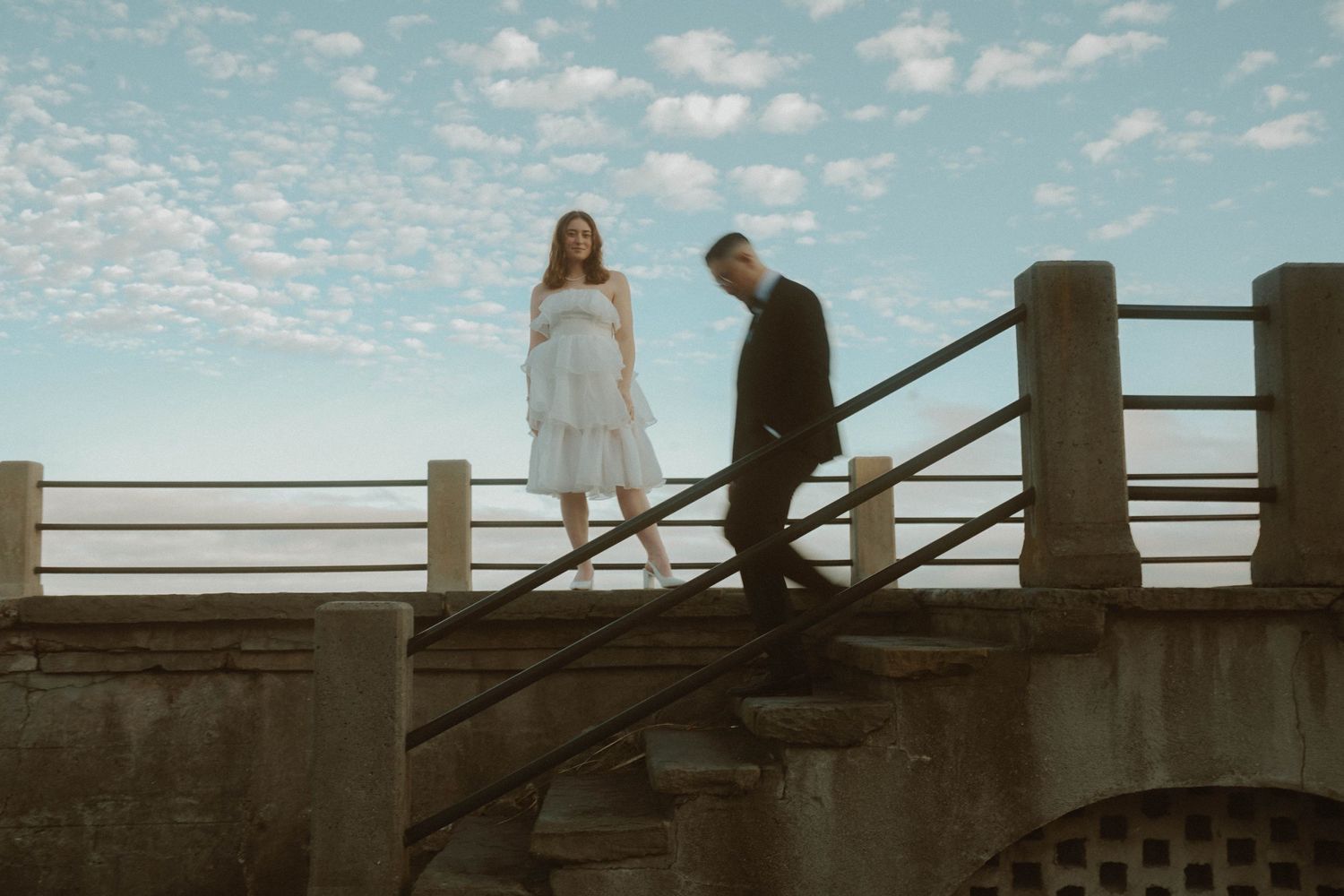A couple in wedding attire climbs stone steps on a bridge under a cloudy blue sky at dusk.