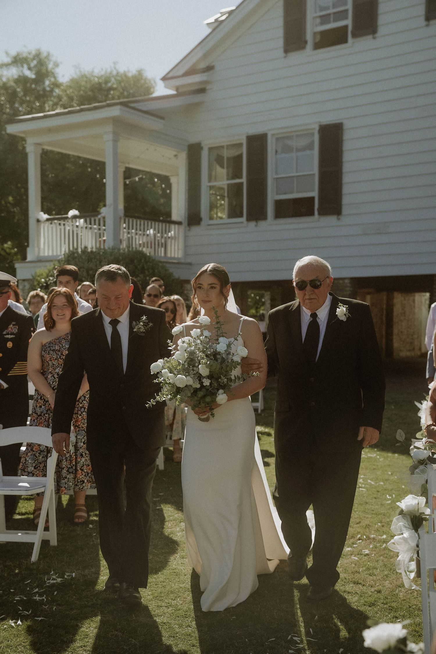 An outdoor wedding ceremony at a white farmhouse with guests lining the walkway and a blooming bouquet.