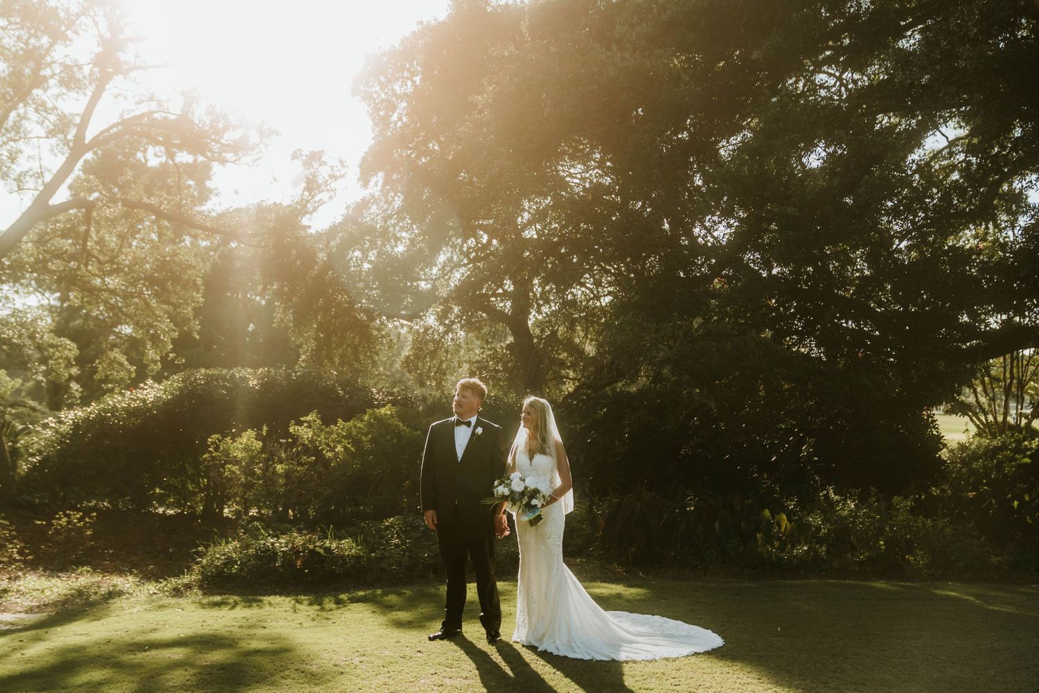 A couple stands in a sunlit garden on their wedding day as golden light streams through the trees.