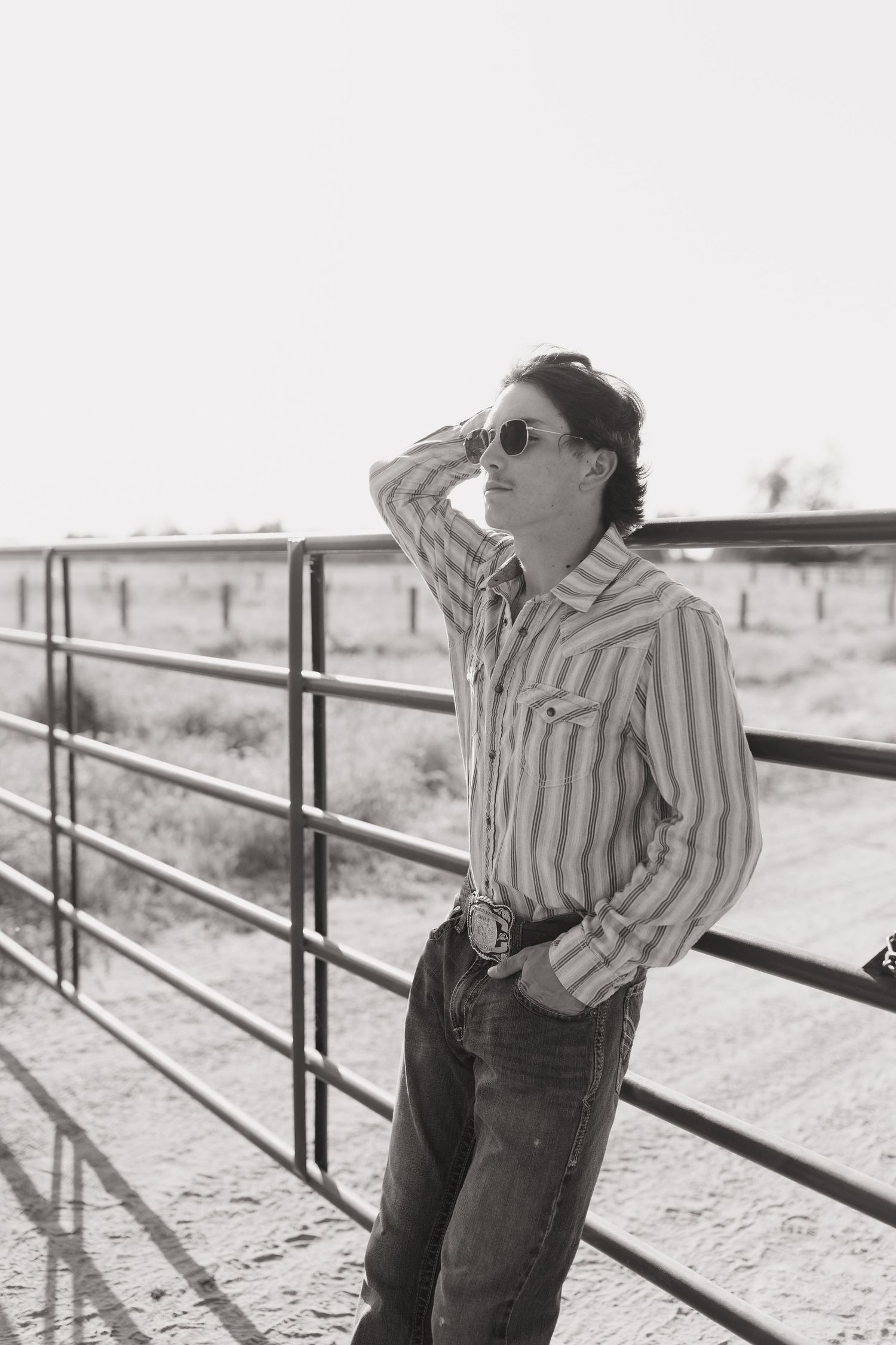 a young man wearing sunglasses out in the midday sun looking out in the distance while leaning against a cattle gate