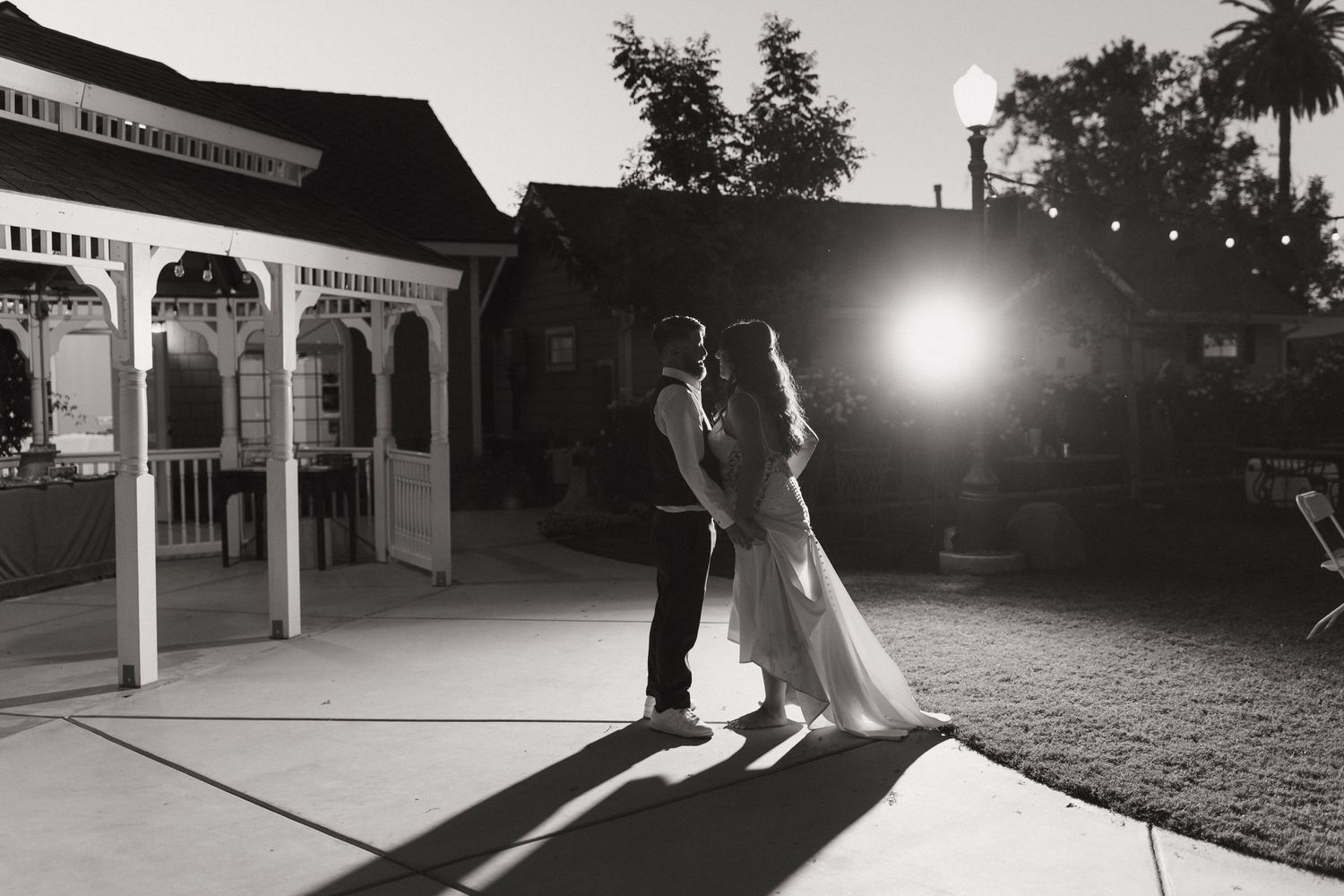 A backlit wedding couple facing each other next to a gazebo in a black and white portrait setting.