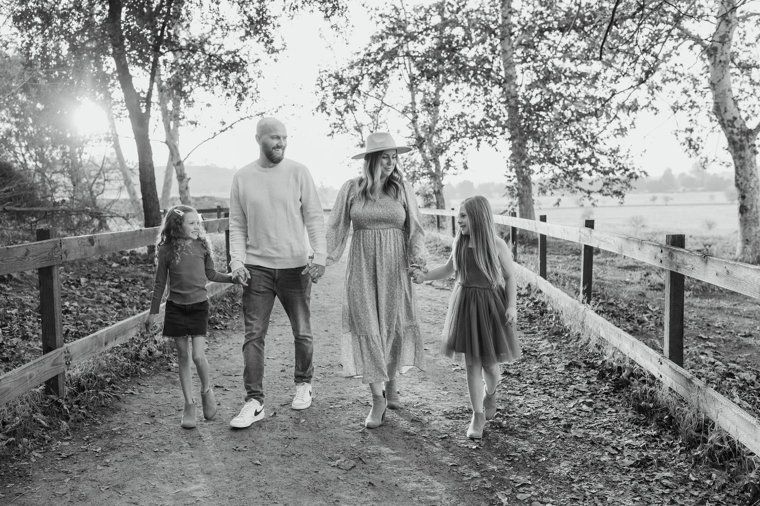 2 adults and 2 children walking together on a tree-lined path with a wooden fence in soft natural light.