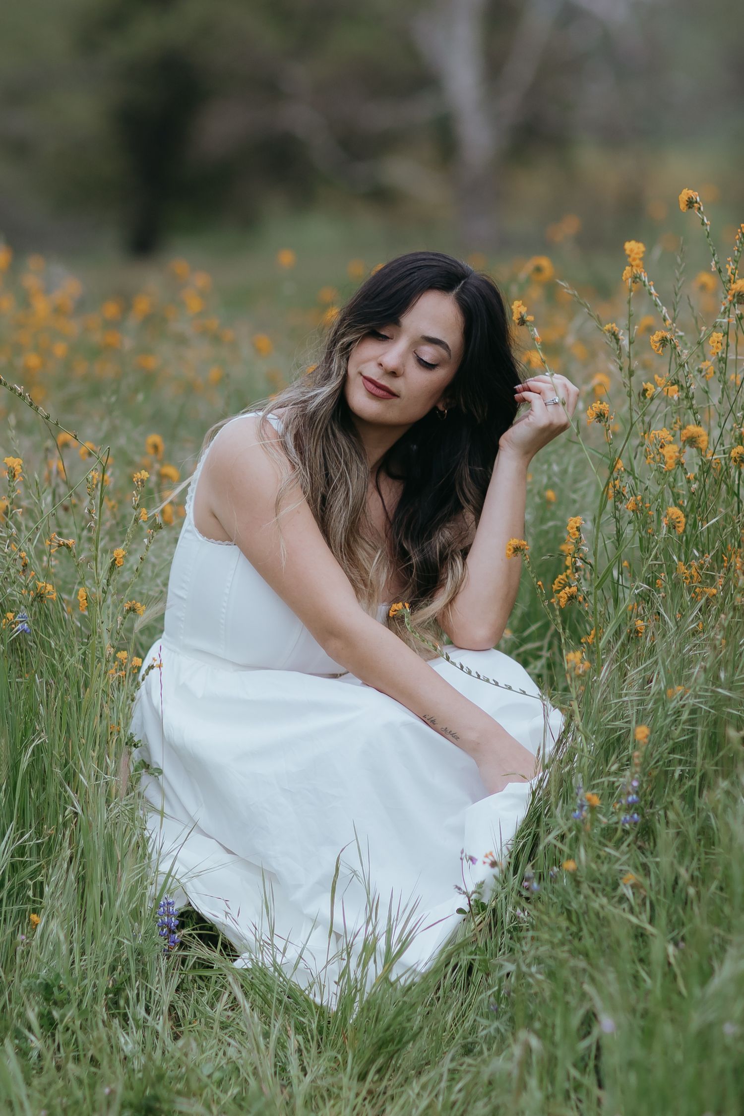 a woman wearing a flowy white dress is softly squatting in a field of yellow wildflowers